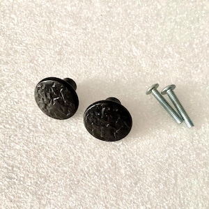May include: Two black, textured cabinet knobs with a hammered metal appearance. The knobs are oval-shaped and have a small, round base. Two silver screws are also included, likely for installation. The hardware is likely for furniture or cabinetry.