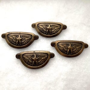 May include: Four semi-circular brass drawer pulls featuring a raised eagle motif with wings spread wide. Each handle has a curved shape with two screw holes on the sides for mounting, showcasing a dark, aged metallic finish with intricate detailing.
