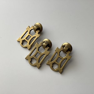 May include: Three antique brass drawer pulls with a decorative, openwork design. Each pull features a rectangular backplate with a stylized, geometric pattern and a rounded knob. The pulls are arranged on a white surface.