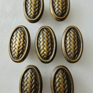 May include: Seven oval cabinet knobs with a woven texture design. The knobs have a bronze finish and are arranged on a white surface. These decorative knobs are suitable for furniture and home decor projects.