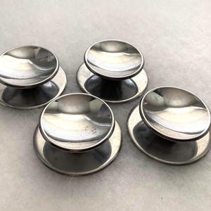 May include: Four circular silver-toned metal knobs with a polished, reflective finish. Each knob features a flat, circular base plate and a raised, concave top section, creating a distinct mushroom-like profile with smooth, curved edges.