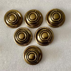 May include: Six gold-toned metal buttons with a concentric circle design. The buttons have a raised center and a smooth, shiny finish.