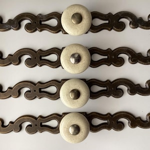 May include: Four antique bronze-toned drawer pulls with cream-colored ceramic knobs. Each pull features an ornate, scrolling design. The knobs have a central metal accent. These decorative pulls are ideal for furniture restoration.