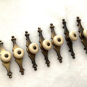 May include: A set of seven drawer pulls, each featuring an elongated, dark brass-toned metal backplate with a pointed, decorative shape and a central, circular off-white ceramic knob accented with a small, dark brown dot in the center.
