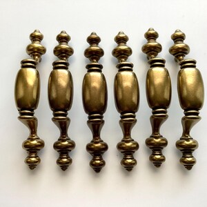 May include: Six antique brass cabinet pulls. Each pull has a decorative design with a cylindrical center and ornate detailing. The pulls are arranged horizontally against a white background.