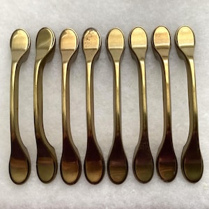 May include: A set of eight matching brass-toned metal cabinet pulls arranged in a vertical row. Each handle features a smooth, elongated, slightly curved body with rounded ends and a polished finish, providing a consistent look for furniture.