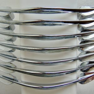 May include: A set of six chrome-plated cabinet pulls. Each pull has a curved, streamlined design with a pointed end on each side. The pulls are arranged in parallel, showcasing their reflective, shiny surface.