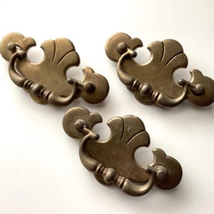 May include: Three antique brass drawer pulls with a decorative, ornate design. Each pull features a curved handle and a stylized, fan-shaped backplate. The pulls are arranged on a white surface, showcasing their aged, vintage appearance.