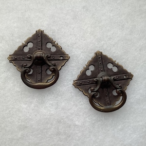 May include: Two matching bronze-toned metal drawer pulls with a diamond-shaped backplate featuring cut-out details and a central vertical bar. Each pull has a hinged, curved drop handle attached to the center, showcasing a dark, aged finish.