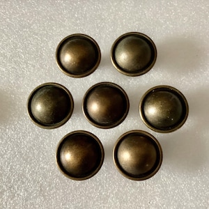 May include: Seven antique bronze-colored furniture tacks arranged in a circular pattern. Each tack has a rounded, domed head and a flat base. The tacks have a slightly weathered appearance, suggesting age or a vintage aesthetic. Ideal for upholstery or decorative projects.