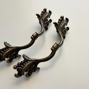 May include: Two antique bronze-toned drawer pulls with ornate, floral-inspired designs. Each pull features a curved handle and decorative flourishes, ideal for adding a vintage touch to furniture. The pulls are set against a white background.