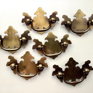 May include: Six antique brass drawer pulls with ornate, symmetrical designs. Each pull features a decorative backplate, a curved handle, and two spherical accents. The brass has a warm, aged patina, suggesting vintage or retro styling.