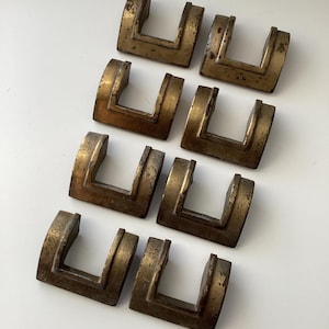 May include: Eight vintage brass drawer pulls in a U-shape design. The pulls have a weathered, aged appearance with a tarnished finish. The hardware is arranged on a white surface, showcasing their unique, antique aesthetic. These are likely for cabinets or drawers.