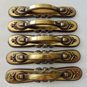 May include: Five antique brass cabinet pulls. Each pull features a curved handle and decorative detailing on a rectangular backplate. The pulls have a warm, aged finish, suggesting vintage or retro design.