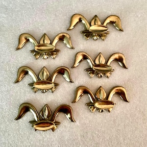 May include: Six antique brass-toned decorative hardware pieces. Each piece features a central oval shape with a stylized floral design and curved, wing-like elements. The hardware is arranged on a textured, off-white surface.