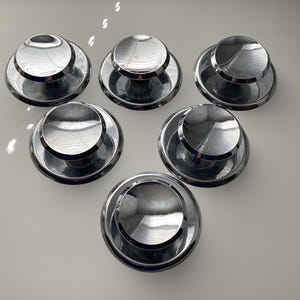 May include: Six shiny, chrome-finished bottle stoppers arranged on a white surface. Each stopper has a circular base and a tiered, domed top. The reflective surfaces create highlights and shadows, giving them a polished appearance. The stoppers are likely for wine or other bottled beverages.