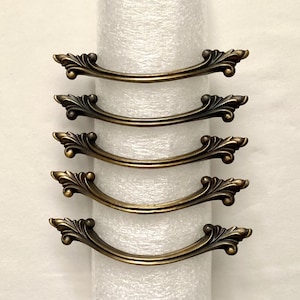 May include: Five antique bronze-colored drawer pulls with a curved handle design. Each pull features ornate, detailed floral accents at each end. The pulls are arranged vertically against a white background.