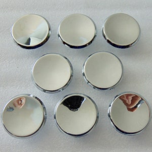 May include: A collection of eight shiny, circular, chrome-plated buttons. Each button has a reflective, convex surface and a polished, metallic rim. The buttons are arranged in a grid-like pattern against a light background.