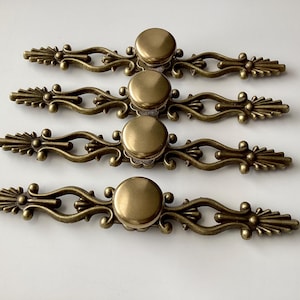 May include: Four antique brass drawer pulls with a decorative design. Each pull features a central round knob and ornate, symmetrical detailing with a vintage aesthetic. The pulls are arranged in parallel on a white surface.