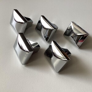May include: Five shiny, chrome-finished cabinet knobs. Each knob has a geometric, diamond-like shape with a rectangular base. The knobs are arranged on a white surface, reflecting light and creating highlights.