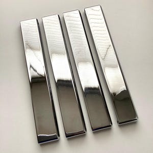 May include: Four rectangular, polished metal bars arranged side by side. The bars have a reflective, mirror-like surface, with a slight beveled edge. The metal appears to be a shiny silver color.