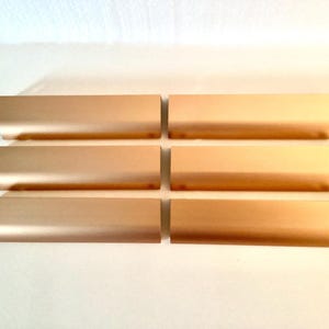 May include: Six rectangular, metallic gold-colored cabinet pulls arranged in two rows of three. The pulls have a curved edge and a smooth, reflective surface. The hardware is likely for furniture or cabinetry.