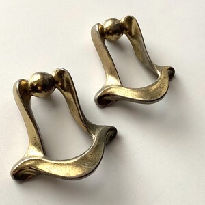 May include: Two vintage brass drawer pulls. Each pull features a curved design with a spherical accent at the top. The pulls have a warm, aged patina, suggesting they are antique or vintage pieces. The design is both functional and decorative.