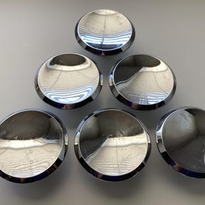 May include: Six shiny, chrome-finished cabinet knobs arranged in a triangular pattern. Each knob has a rounded, domed shape with a reflective surface, and a polished, circular rim. The knobs are set against a light gray background.