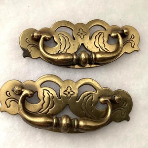 May include: Two brass drawer pulls featuring an ornate, openwork backplate with engraved leaf motifs and a central four-petaled flower design. Each pull includes a curved, swinging handle attached to the backplate with two metal mounting points.