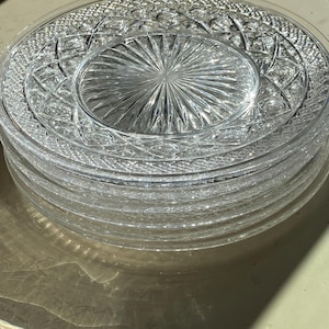 May include: A stack of clear glass plates featuring a central sunburst pattern surrounded by a wide border of intricate diamond and cross-hatch geometric textures. The plates have a smooth, raised outer rim and a reflective, transparent finish.