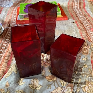 May include: Three red glass vases of varying sizes and shapes, arranged on a floral patterned tablecloth.