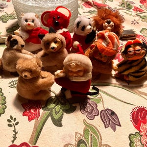 May include: A collection of vintage plush animal toys, including a panda, koala, raccoon, bear, lion, and Garfield. The toys are in various colors, including brown, red, white, and orange. The toys are arranged on a floral patterned tablecloth.