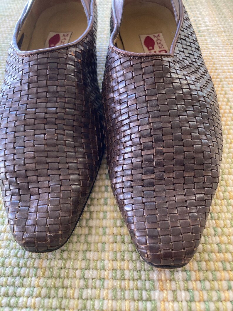 Puede incluir: Primer plano de un par de zapatos de cuero marr&oacute;n tejido. Los zapatos tienen un dise&ntilde;o sin cordones con punta puntiaguda. El patr&oacute;n tejido cubre todo el zapato, creando una apariencia texturizada. El interior del zapato es visible, con una etiqueta.