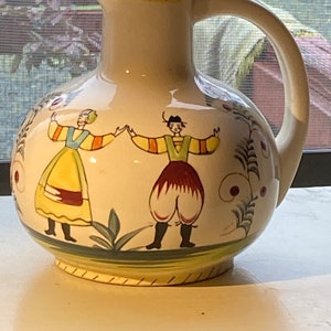 May include: Cream-colored ceramic pitcher with a rounded body and a curved handle. The pitcher features a painted design of two figures in traditional clothing, holding hands. The rim and base are accented with green and yellow trim. Decorative floral elements are also present.
