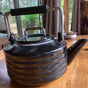 May include: Black stovetop kettle with a ribbed design. The kettle has a black handle and spout, with a silver metal handle and lid handle. The kettle is sitting on a wooden surface.