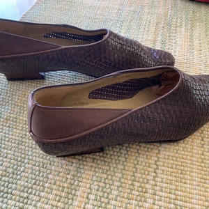 Puede incluir: Un par de zapatos de cuero marr&oacute;n tejido con punta y tac&oacute;n bajo. Los zapatos tienen un patr&oacute;n tejido en la parte superior y un ribete de cuero marr&oacute;n liso. Los zapatos se exhiben sobre una superficie estampada en verde claro y blanco.