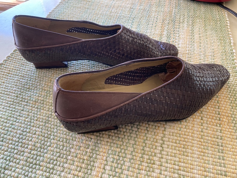 Puede incluir: Un par de zapatos de cuero marr&oacute;n tejido con punta y tac&oacute;n bajo. Los zapatos tienen un patr&oacute;n tejido en la parte superior y un ribete de cuero marr&oacute;n liso. Los zapatos se exhiben sobre una superficie estampada en verde claro y blanco.