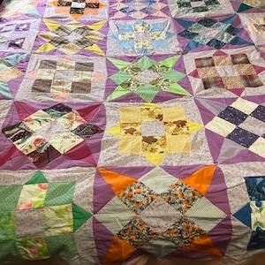 May include: A patchwork quilt featuring a variety of square blocks in diverse colors and patterns, including floral, plaid, and solid fabrics. The blocks are separated by light purple sashing and arranged in a grid pattern with star-shaped motifs.