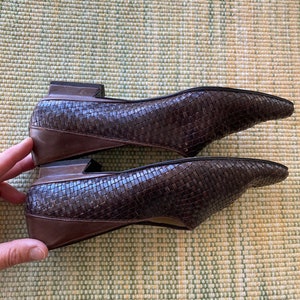 Puede incluir: Un par de bailarinas de cuero marr&oacute;n tejido con puntera. Los zapatos tienen un tac&oacute;n bajo y un ribete marr&oacute;n oscuro. Los zapatos se exhiben sobre una superficie estampada en verde claro y amarillo.