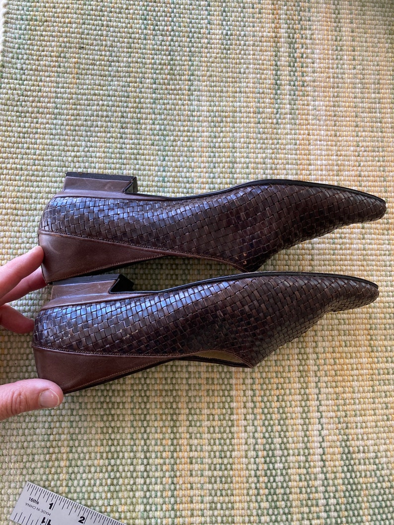 Puede incluir: Un par de bailarinas de cuero marr&oacute;n tejido con puntera. Los zapatos tienen un tac&oacute;n bajo y un ribete marr&oacute;n oscuro. Los zapatos se exhiben sobre una superficie estampada en verde claro y amarillo.
