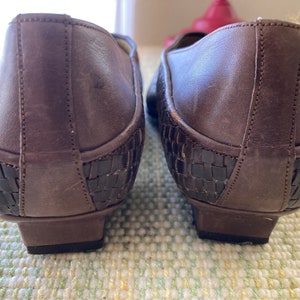 Puede incluir: Un par de zapatos de cuero marr&oacute;n con un dise&ntilde;o tejido en el tal&oacute;n. Los zapatos tienen un tac&oacute;n bajo y una punta redondeada. El cuero parece envejecido, d&aacute;ndoles un aspecto vintage. Zapatos para uso diario.