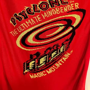 May include: Red t-shirt featuring the text "Psyclone The Ultimate Mindbender" in black and yellow, with a graphic of a roller coaster and the words "Magic Mountain".
