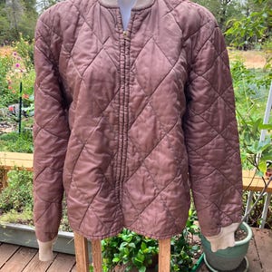 May include: A mauve quilted bomber jacket featuring a diamond-stitched pattern, a full-length front zipper, and ribbed knit collar and cuffs in a light beige tone. The jacket has long sleeves and a standard fit, displayed on a mannequin torso.
