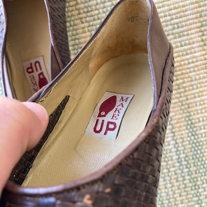 Puede incluir: Primer plano de un par de bailarinas de cuero marr&oacute;n tejido. El interior del zapato presenta una plantilla color crema con el texto "MAKE UP" impreso en una etiqueta roja. Los zapatos parecen sin usar.