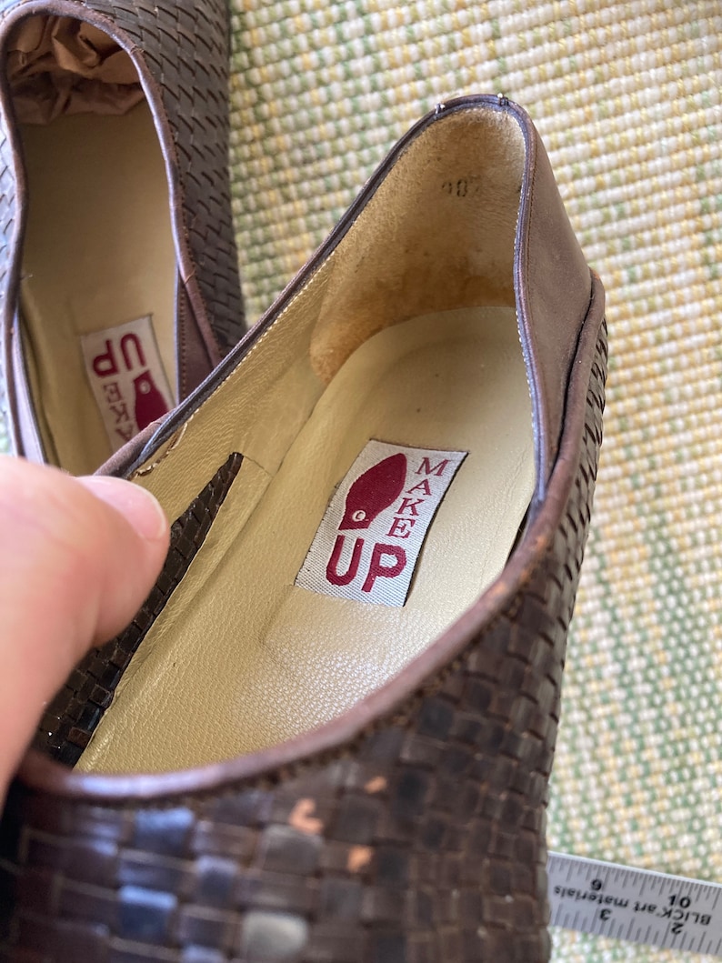 Puede incluir: Primer plano de un par de bailarinas de cuero marr&oacute;n tejido. El interior del zapato presenta una plantilla color crema con el texto "MAKE UP" impreso en una etiqueta roja. Los zapatos parecen sin usar.