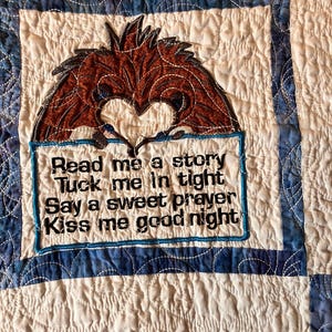 May include: A white quilted fabric with a blue border and a brown embroidered bird. The bird is holding a sign that says "Read me a story, Tuck me in tight, Say a sweet prayer, Kiss me good night."