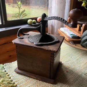 May include: An antique coffee grinder with a dark wooden box and a cast iron grinding mechanism. The grinder has a crank handle and a decorative base. The wood has a weathered appearance, suggesting age and use. The grinder sits on a woven rug.