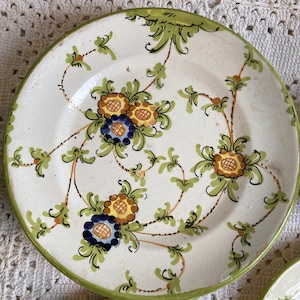 May include: White ceramic plate with a green rim and floral design. The plate features a pattern of green leaves and brown stems with yellow and blue flower accents. The plate is part of a set.