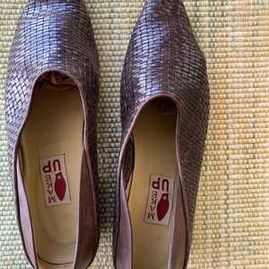 Puede incluir: Un par de zapatos de cuero trenzado marr&oacute;n. Los zapatos tienen punta y un dise&ntilde;o sin cordones. Las plantillas tienen una etiqueta que dice "MAKE UP" con un gr&aacute;fico rojo. Los zapatos se exhiben sobre una superficie texturizada.