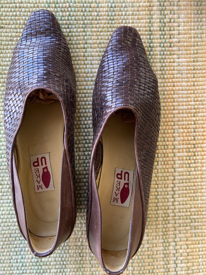 Puede incluir: Un par de zapatos de cuero trenzado marr&oacute;n. Los zapatos tienen punta y un dise&ntilde;o sin cordones. Las plantillas tienen una etiqueta que dice "MAKE UP" con un gr&aacute;fico rojo. Los zapatos se exhiben sobre una superficie texturizada.